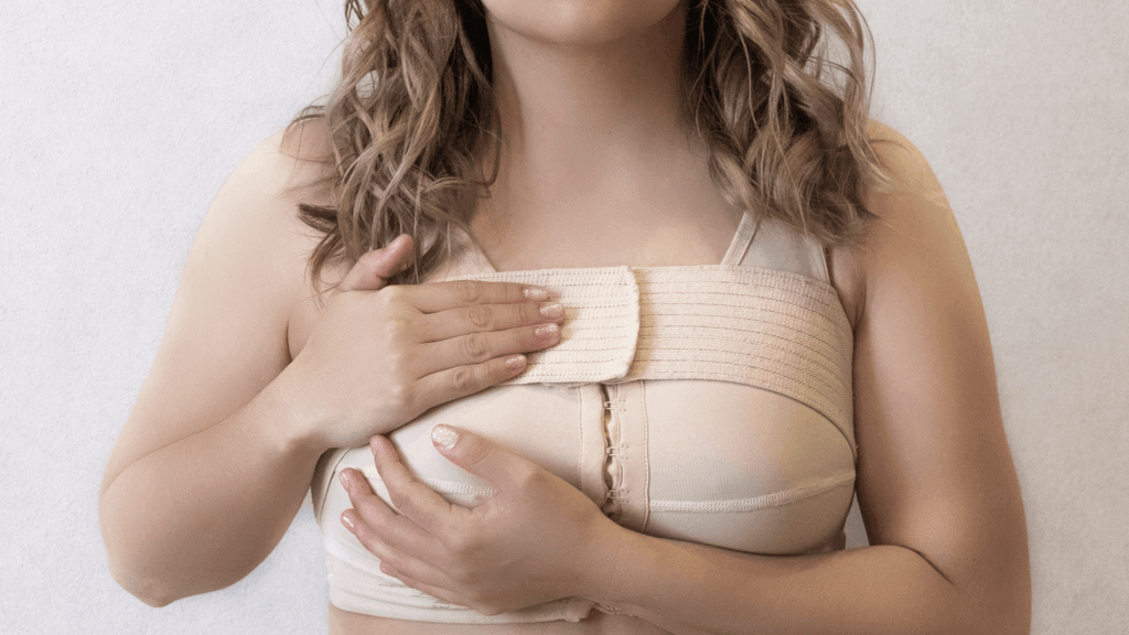 A woman wearing a beige post-surgical compression bra, an essential step in managing breast lymphedema after cancer treatment, holds her chest with both hands. Only the lower part of her face and torso are visible.