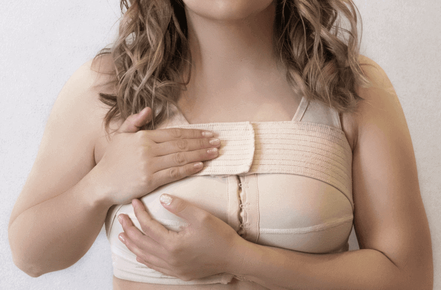 A woman wearing a beige post-surgical compression bra, an essential step in managing breast lymphedema after cancer treatment, holds her chest with both hands. Only the lower part of her face and torso are visible.