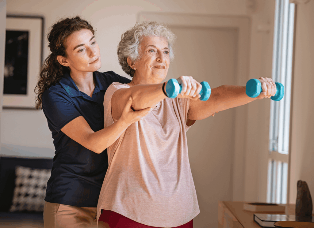 A young caregiver assists an older woman with gray hair as she lifts small blue dumbbells