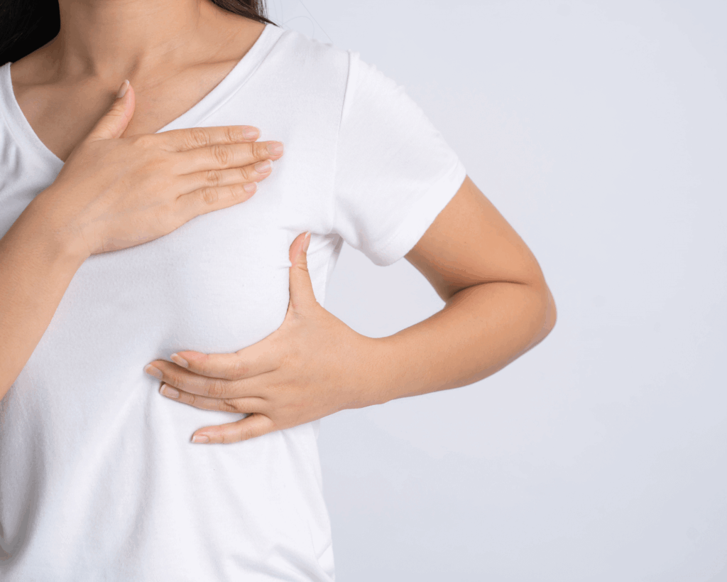 A person in a white t-shirt performs a breast self-exam, checking for signs like swelling that could indicate breast cancer. One hand feels the breast while the other rests on their upper chest against a plain, light background.