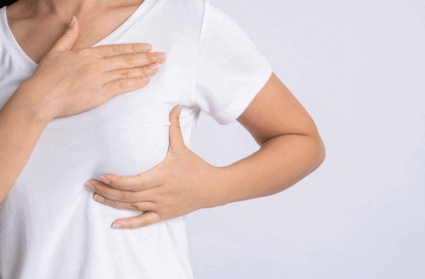 A person in a white t-shirt performs a breast self-exam, checking for signs like swelling that could indicate breast cancer. One hand feels the breast while the other rests on their upper chest against a plain, light background.