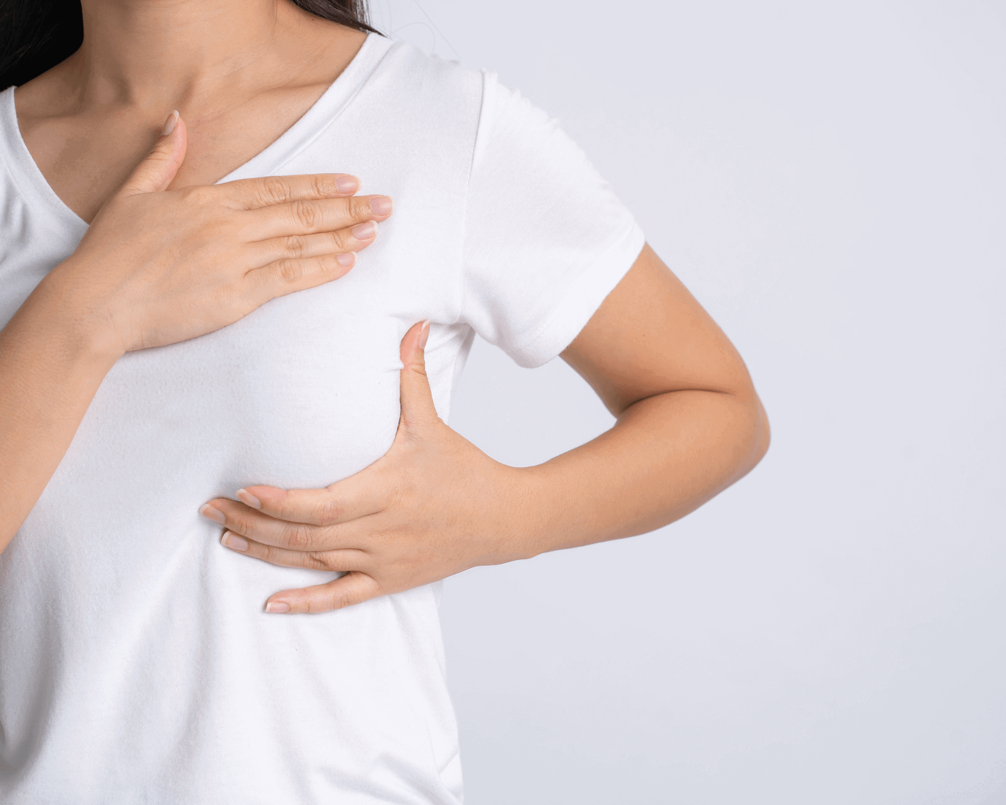 A person in a white t-shirt performs a breast self-exam, checking for signs like swelling that could indicate breast cancer. One hand feels the breast while the other rests on their upper chest against a plain, light background.