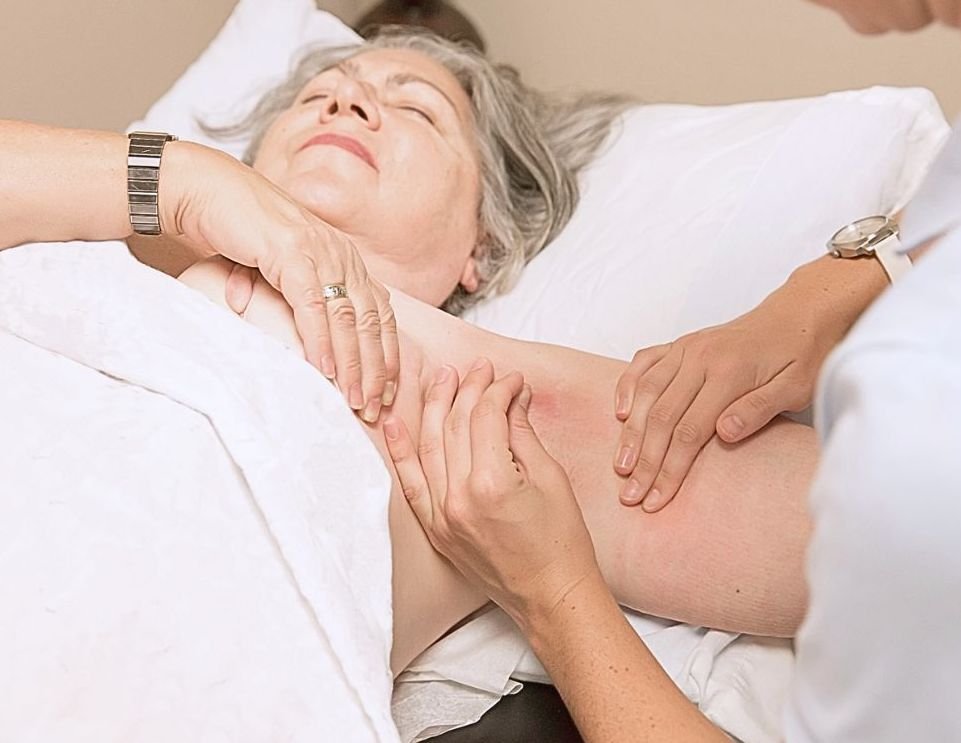 older woman laying on white pillow covered with white sheet getting manual physical therapy done by a therapist to her axilla and shoulder
