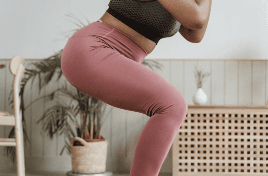 woman in pink leggings and black sports bra doing a squat in her living room with a plant and woven table behind her