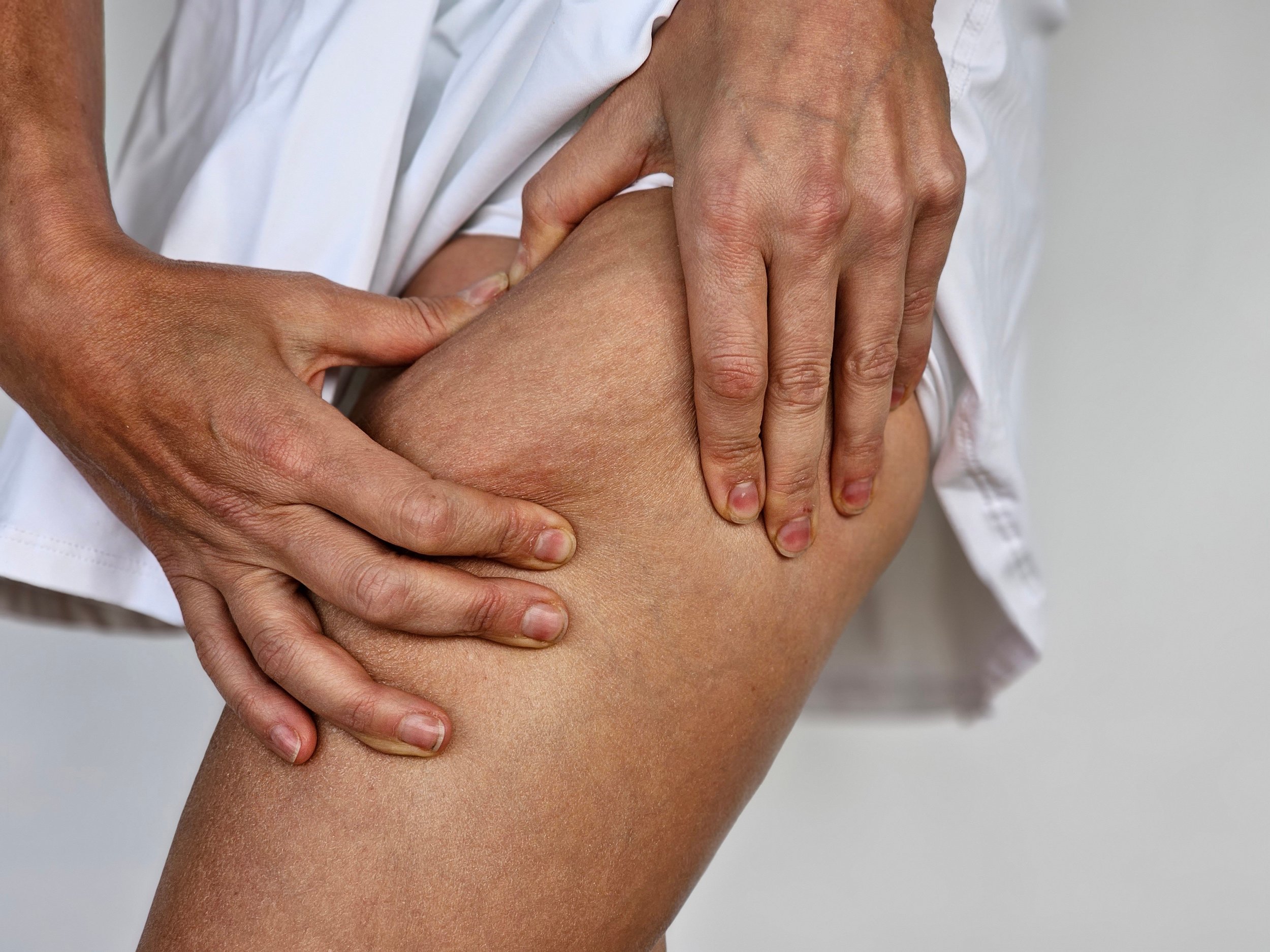 A person wearing a white garment is pressing and examining the skin on their upper thigh with both hands, possibly checking for left thigh swelling or other related issues.