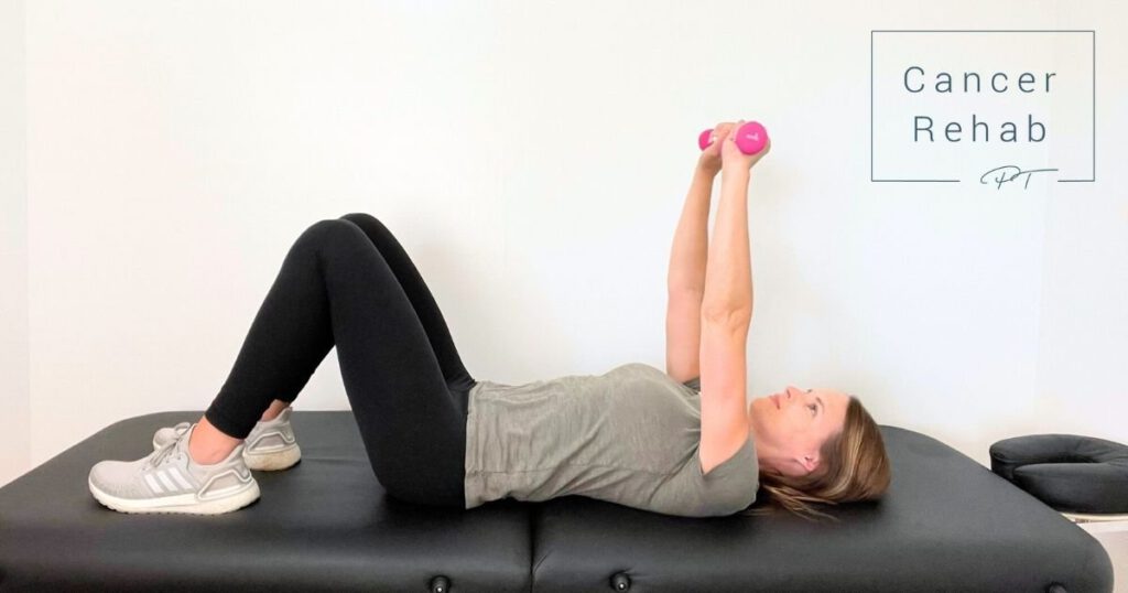 A physical therapist lies on her back on a black exercise table, knees bent, holding pink dumbbells with both hands extended above her chest, highlighting exercises for breast lymphedema.