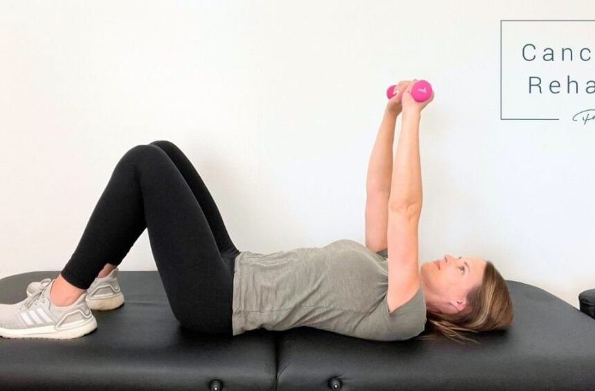 A physical therapist lies on her back on a black exercise table, knees bent, holding pink dumbbells with both hands extended above her chest, highlighting exercises for breast lymphedema.
