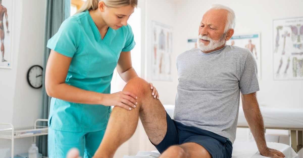 A healthcare professional examines an elderly man's knee in a medical office, checking for ways to reduce fluid for knee swelling as the man watches during the gentle assessment.