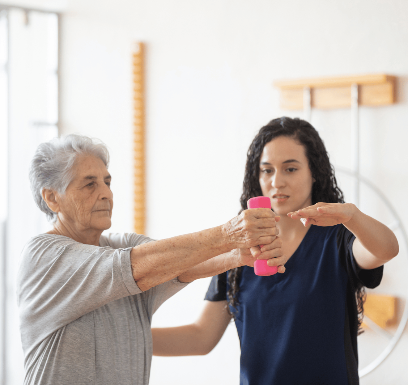Show thumbnail preview
An elderly woman holds a pink dumbbell with both hands while a female therapist guides her arm movement during a physical therapy session in a bright room.