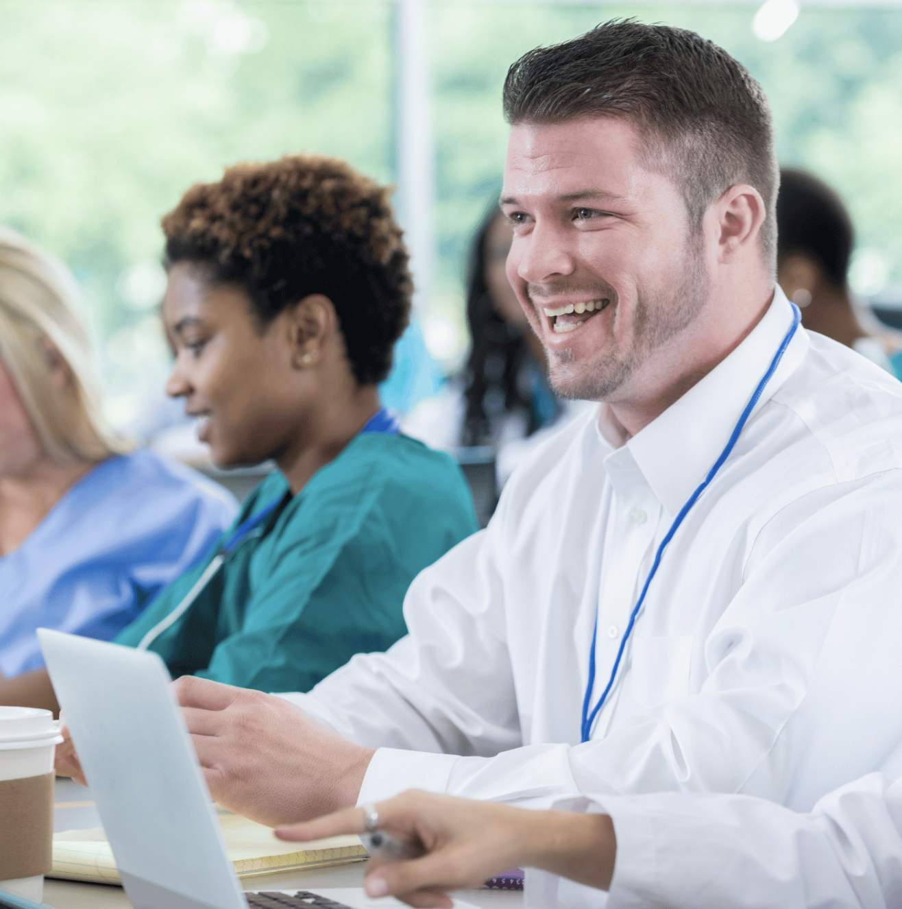 Show thumbnail preview
A group of people in professional attire, including medical scrubs, sit at a table with laptops, papers, and coffee. One man in a white shirt is smiling and engaged in conversation. The setting appears to be a meeting or training session.