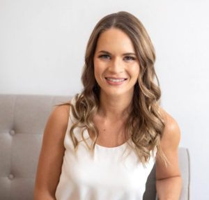 Kelly Sturm, a specialist in cancer and lymphedema physical therapy with long wavy hair, sits on a light-colored sofa in a sleeveless white top and smiles at the camera against a plain white background.