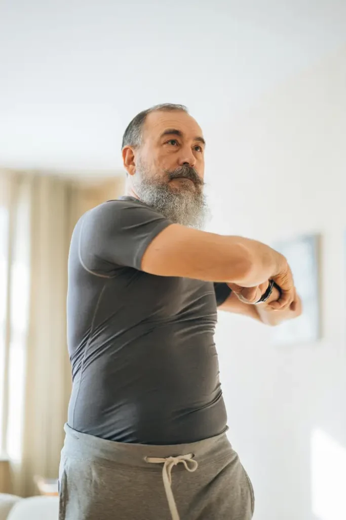 male with gray hair and long beard, wearing gray shirt, doing stretches at home, as instructed by his lymphedema physical therapist