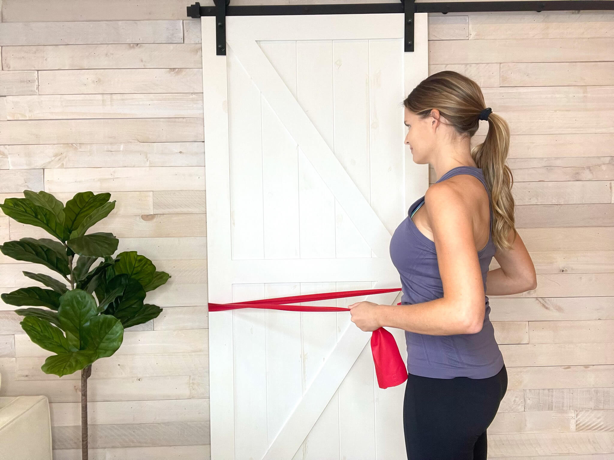 A woman in workout clothes stands sideways, pulling a red resistance band anchored to a sliding barn door, exercising her arm and back muscles. She is doing physical therapy for shoulder pain.