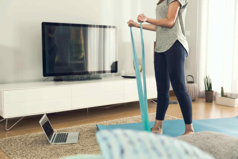 A woman stands on a yoga mat at home, holding a resistance band, facing a laptop on the floor