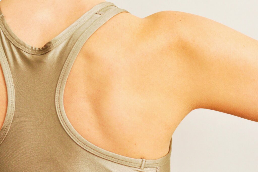 A close-up of a persons upper back and shoulder wearing a beige sleeveless top, with one arm slightly raised, showing smooth skin and defined shoulder blade