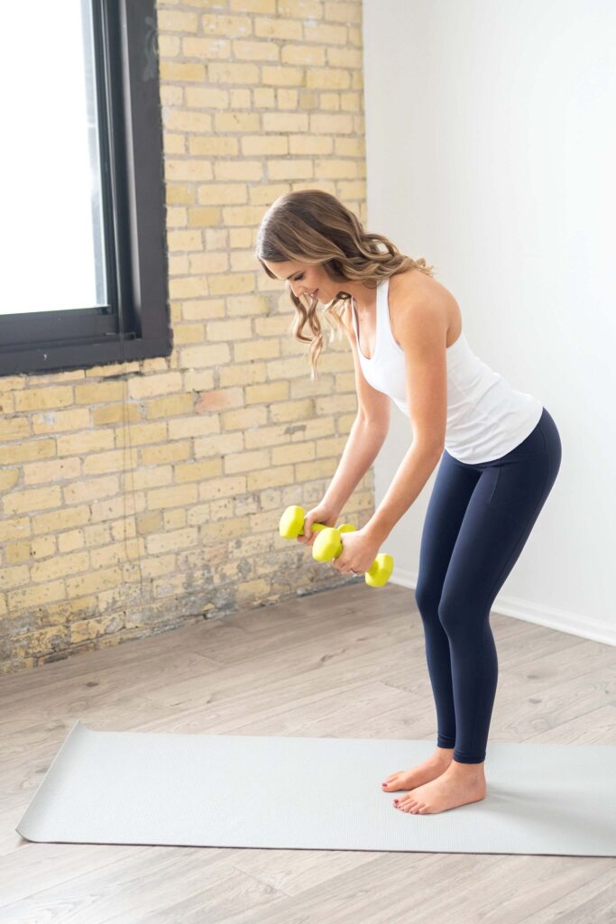 A woman in workout clothes stands on a yoga mat indoors, holding yellow dumbbells and bending forward at the hips, performing an exercise, part of physical therapy for shoulder pain. She is in a bright room with a brick wall and a large window.
