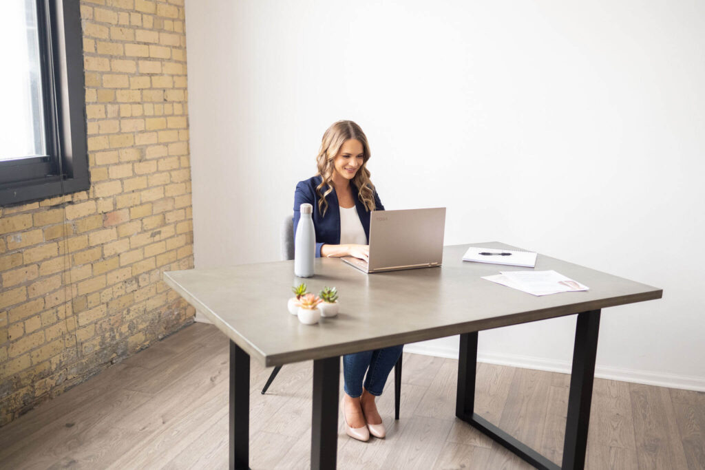A woman with long, wavy hair sits at a desk working on a laptop. The desk also has a water bottle, small potted plants, papers, and a notebook. The background is plain white.