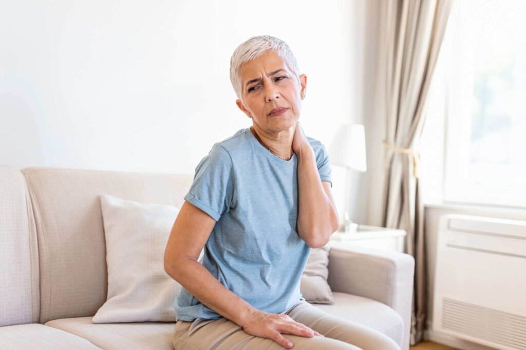 Older woman with short gray hair sitting on a couch, holding her neck with a pained expression, suggesting discomfort or neck pain. She is wearing a light blue shirt and beige pants in a bright living room.