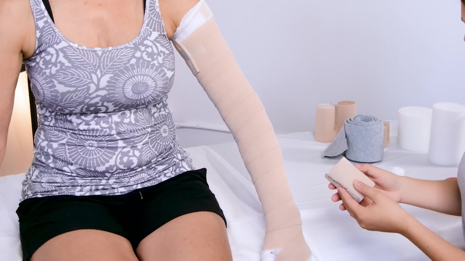 A person wearing a patterned tank top and black shorts sits on an exam table with their left arm fully wrapped in a bandage as part of lymphedema treatment, while another person prepares a roll of bandage nearby.