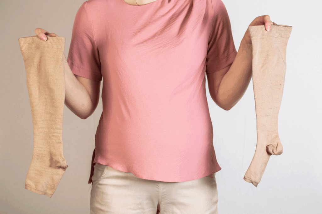 A person wearing a pink shirt and beige pants holds up a pair of beige compression stockings, one in each hand, against a plain light background.