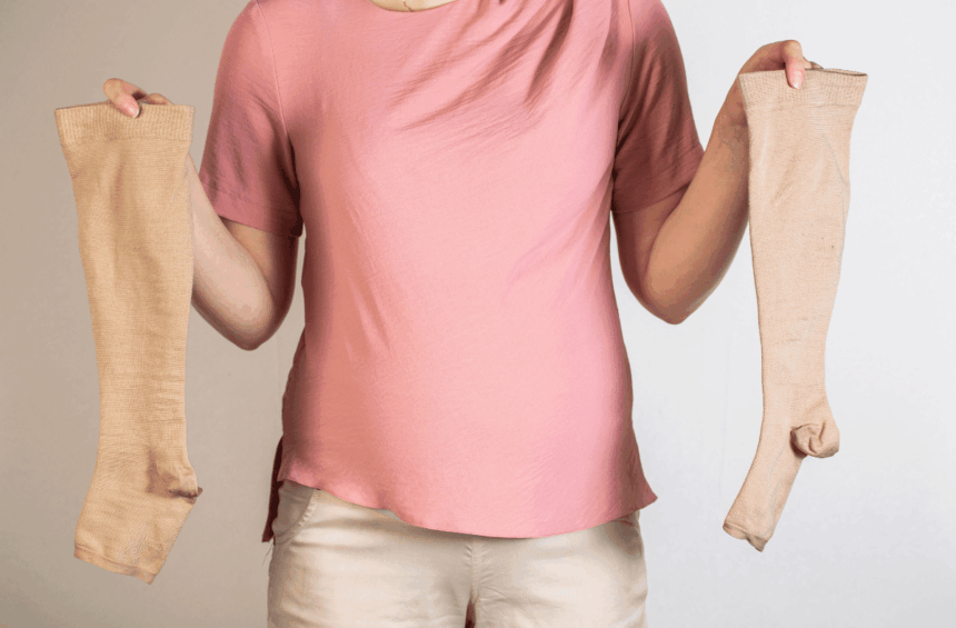 A person wearing a pink shirt and beige pants holds up a pair of beige compression stockings, one in each hand, against a plain light background.