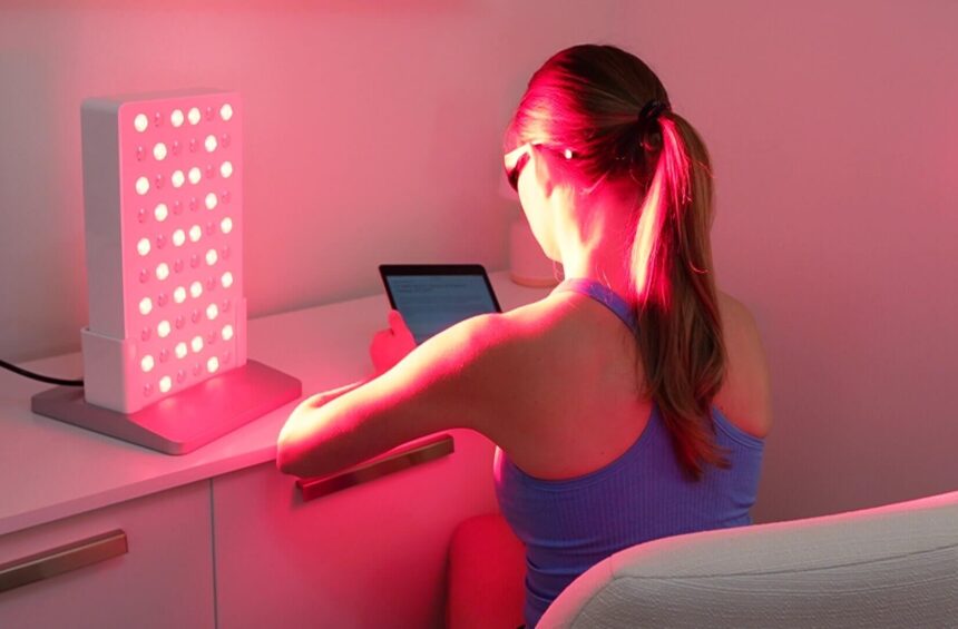 physical therapist sitting in red light therapy, relaxing and getting some work done on ipad with red light panel turned on next to her