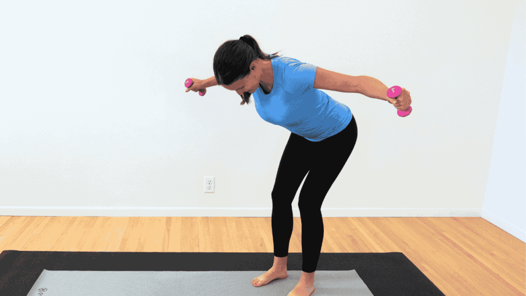 physical therapist in blue shirt and black pants on gray yoga mat, standing bent over with arms out to the side, hold smalling, yellow dumbbells showing exercise for breast cancer survivors