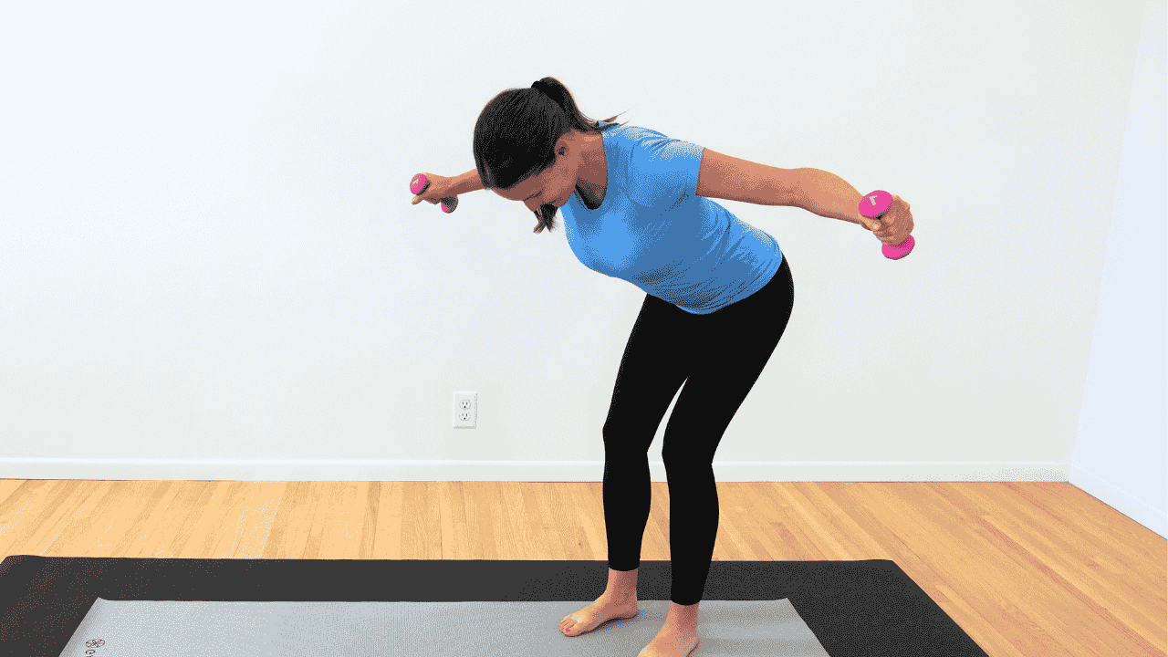 physical therapist in blue shirt and black pants on gray yoga mat, standing bent over with arms out to the side, hold smalling, yellow dumbbells showing exercise for breast cancer survivors