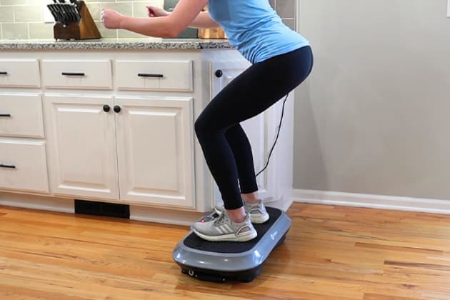A woman standing on a vibration plate in a neutral squat position with arms extended out in front of her. 