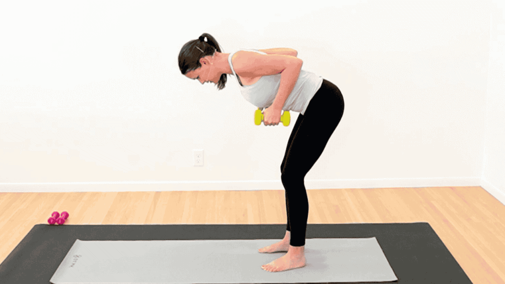 physical therapist in workout clothes, standing bent over on a yoga mat. Holding yellow dumbbells and rowing them up to her sides to show exercise after breast cancer radiation
