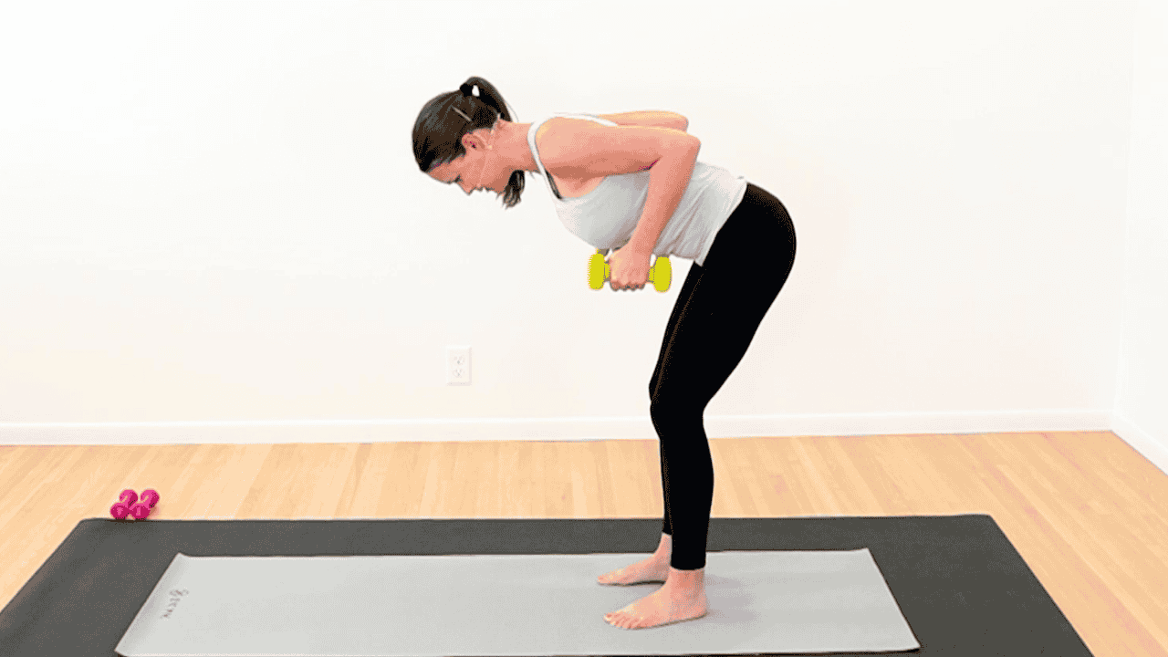 physical therapist in workout clothes, standing bent over on a yoga mat. Holding yellow dumbbells and rowing them up to her sides to show exercise after breast cancer radiation