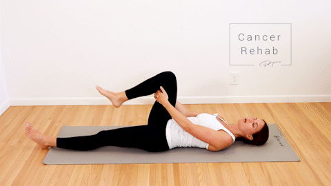A certified lymphedema therapist lying on her back with one leg straight down and one leg bending up and down, stretching her hamstring.
