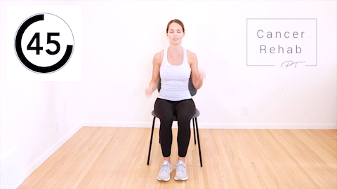 A woman sitting in a chair and extending her arm overhead into a shoulder press as she kicks her leg out.