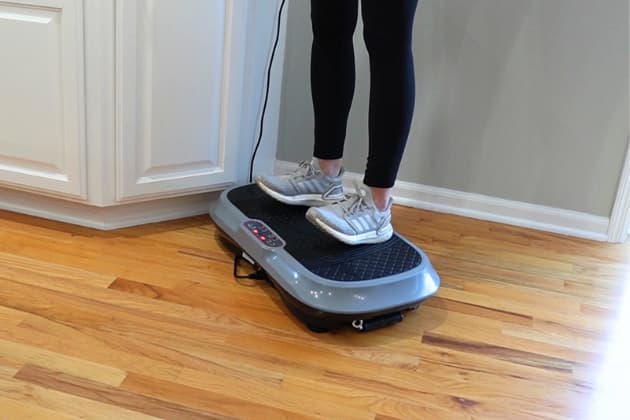 A woman standing on a vibration plate, lifting her toes off the plate and balancing on her heels.