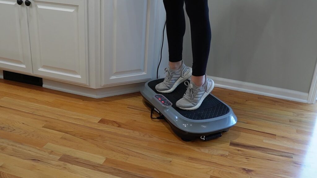 A woman standing on a vibration plate, lifting her heels off the plate and balancing on her toes.