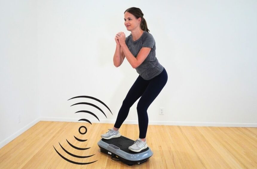physical therapist in gray shirt standing on vibration plate doing a side lunge in a room with white walls