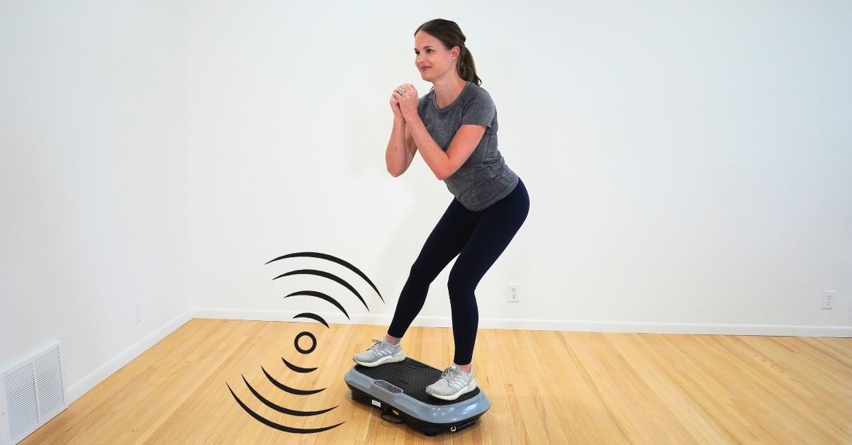 physical therapist in gray shirt standing on vibration plate doing a side lunge in a room with white walls